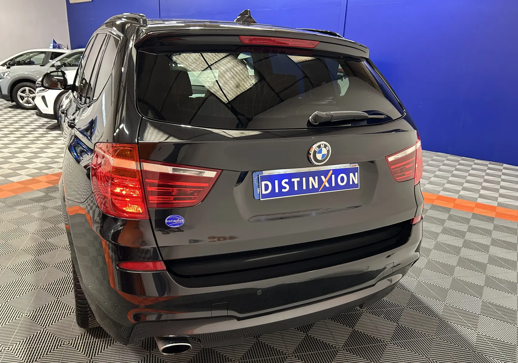 Vue 3/4 arrière d’un BMW X3 noir avec attelage, feu arrière allumé, dans un showroom au sol à damier.