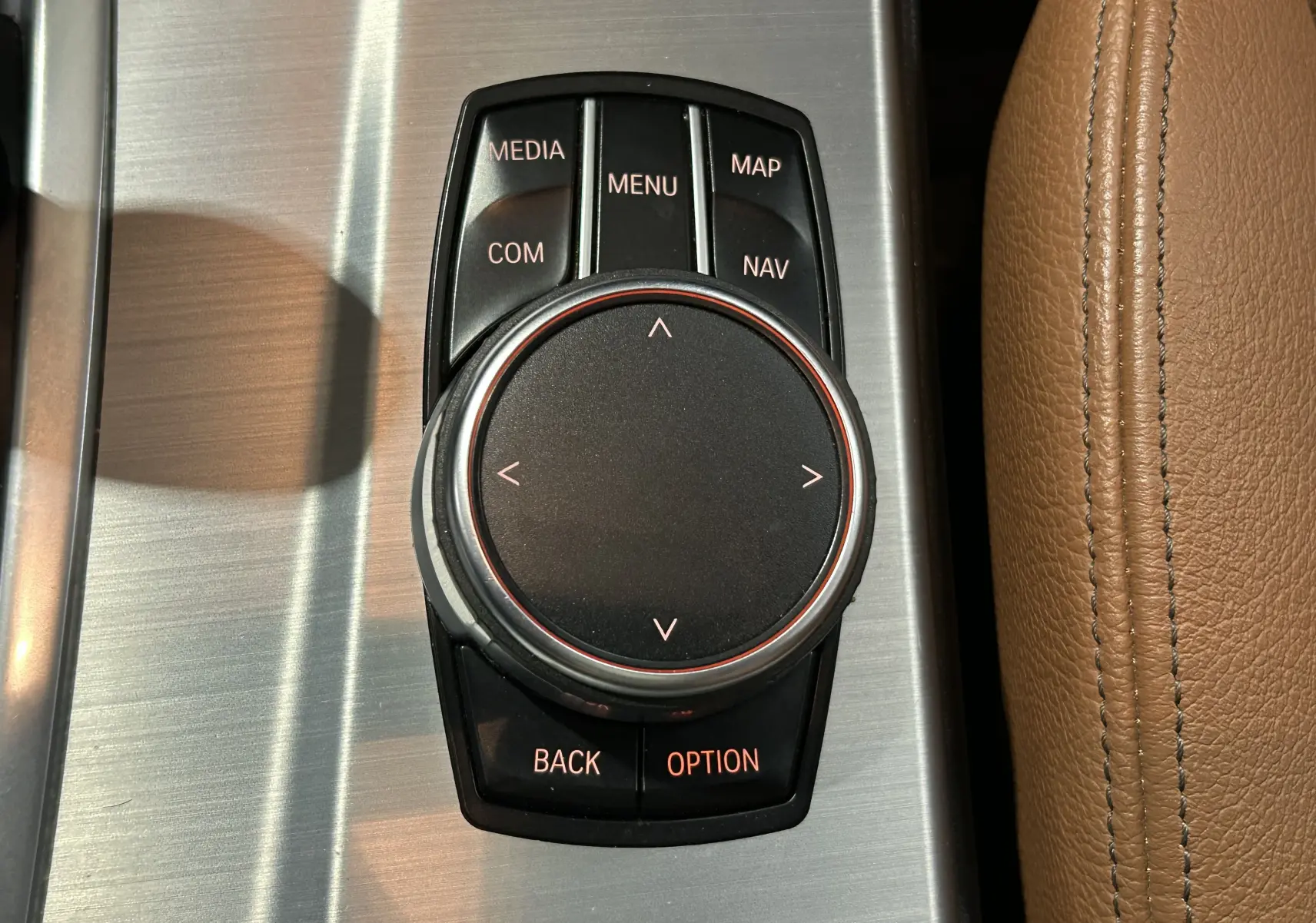 Gros plan sur la molette de commande centrale noire avec boutons autour, sur console en aluminium brossé et cuir marron.