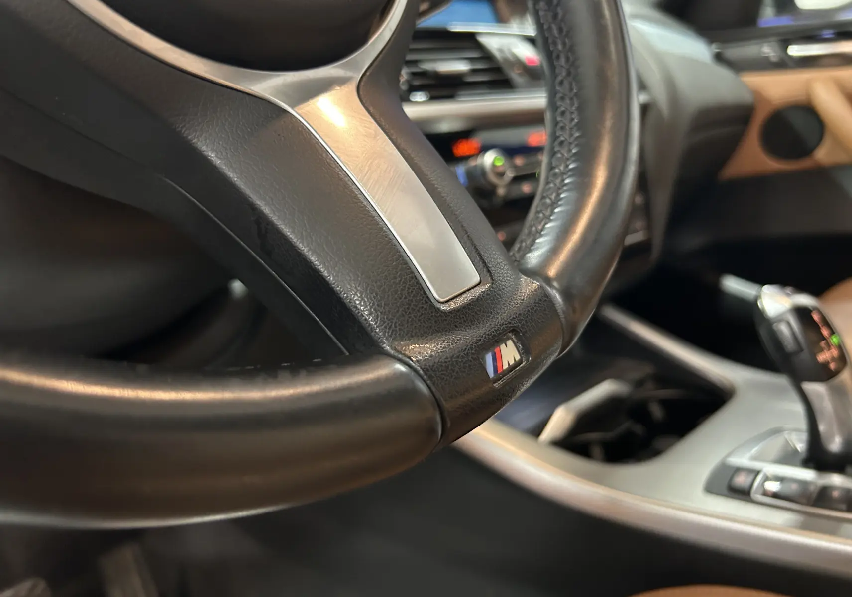 Gros plan sur le volant noir avec logo M et levier de vitesses dans l’habitacle marron d’une BMW X3 sDrive18dA 2017.