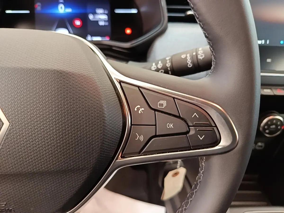 Gros plan sur le volant cuir synthétique de la Renault Clio gris clair, avec commandes multifonctions et tableau de bord numérique flou.
