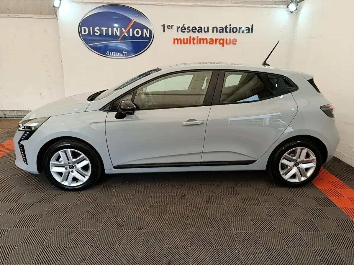 Profil côté gauche d'une Renault Clio gris clair 2025, avec jantes alliage et rétroviseurs noirs, en intérieur showroom.