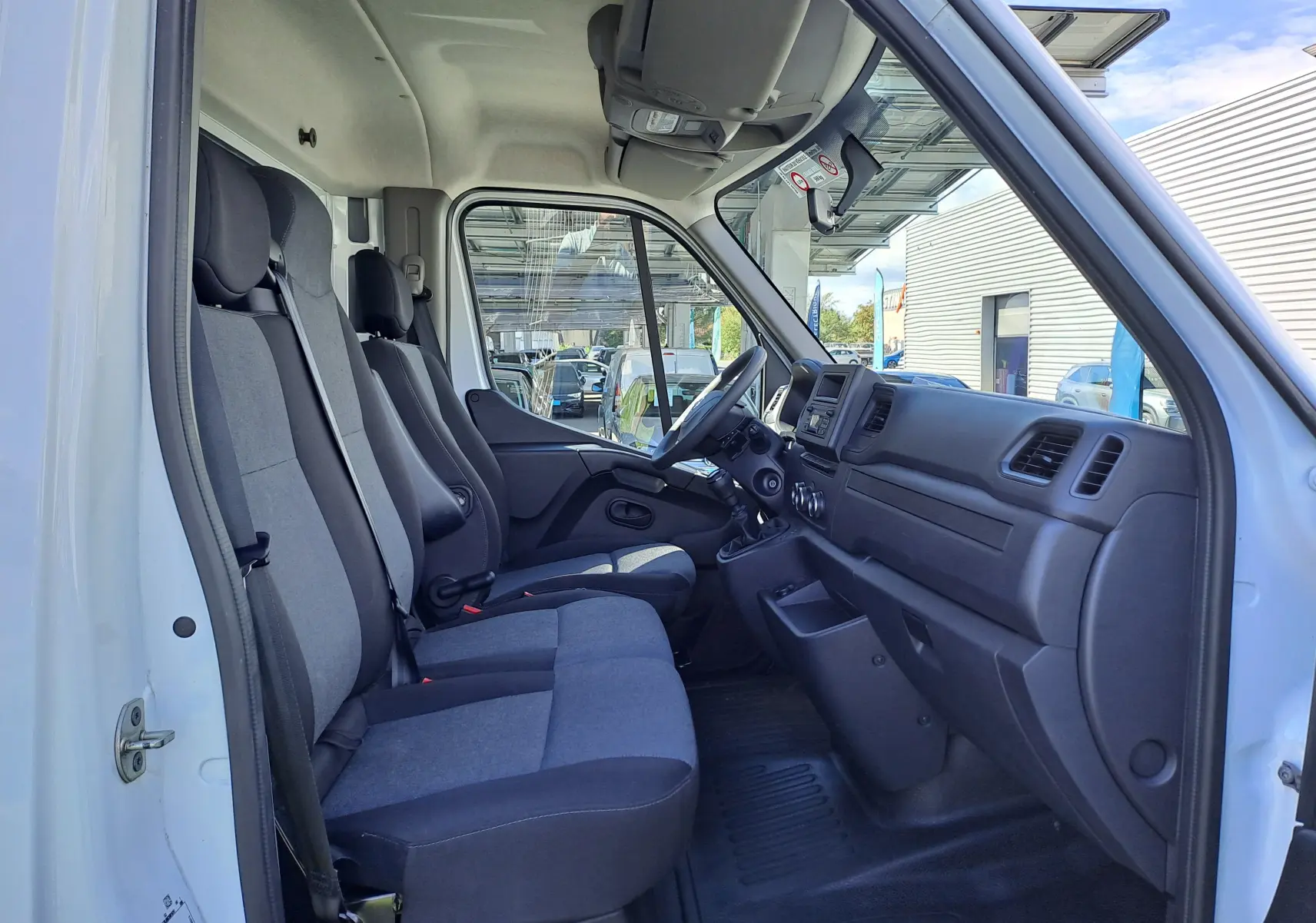 Vue intérieure côté conducteur du Nissan Interstar blanc 2024, montrant les sièges en tissu gris et le tableau de bord fonctionnel.