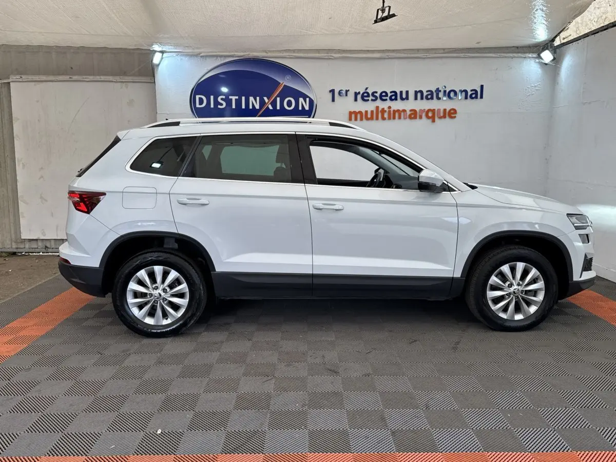 Vue latérale droite d'un Skoda Karoq blanc 2024 avec jantes alliage et barres de toit noires dans un showroom.