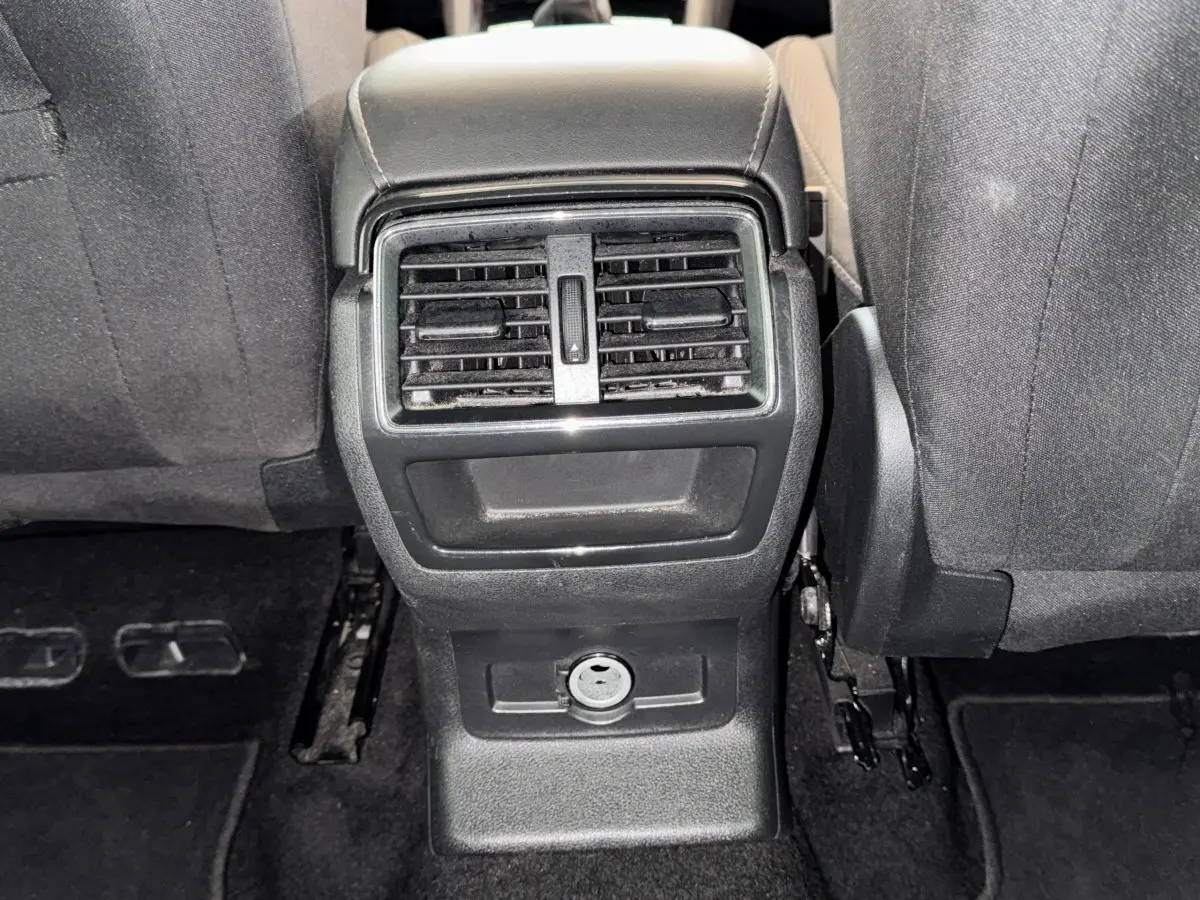 Vue rapprochée de la console centrale arrière noire avec bouches d’aération et prise 12V entre les sièges avant gris foncé du Skoda Karoq blanc.