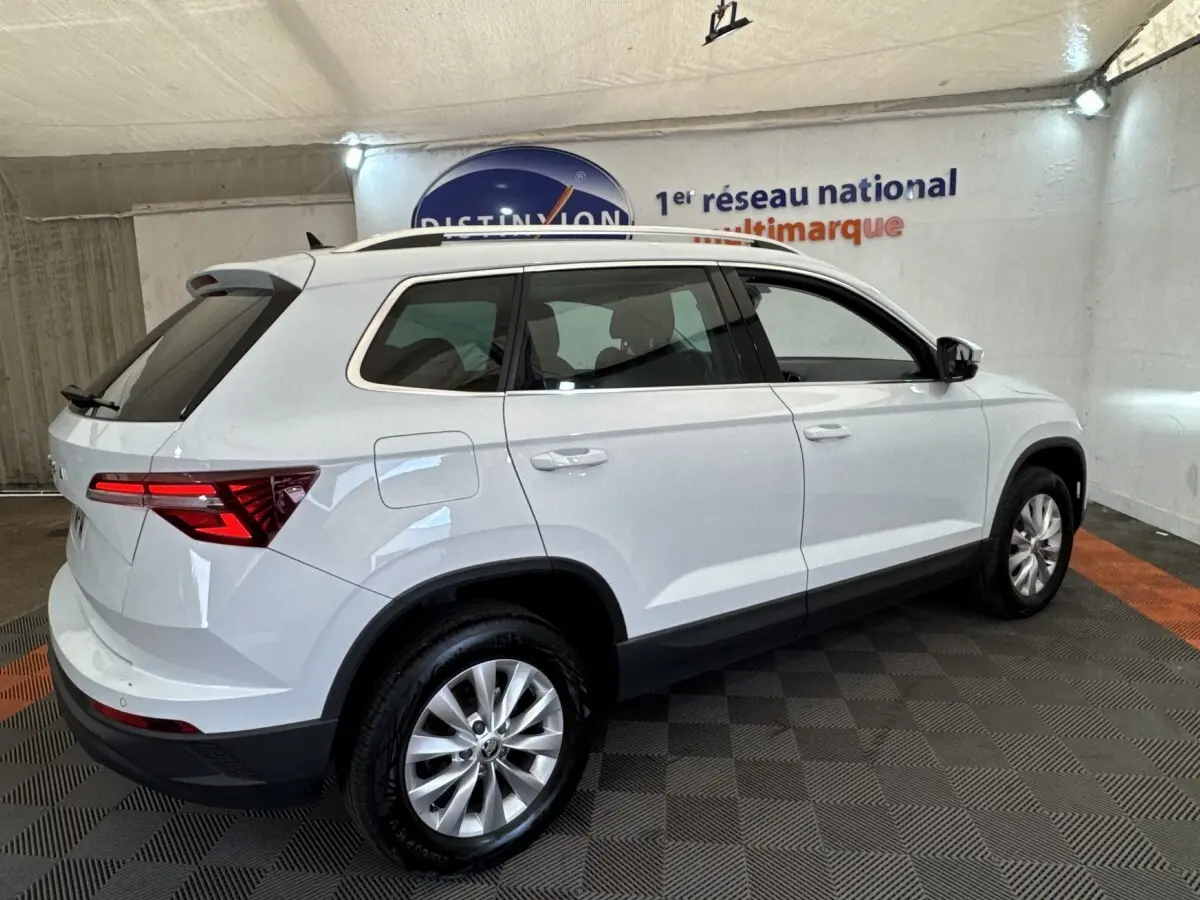Vue 3/4 arrière droite d’un Skoda Karoq blanc 2024 dans un showroom, avec feux arrière LED et barres de toit noires.