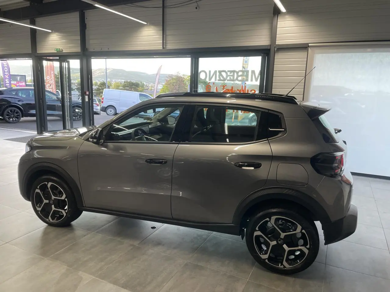 Vue de profil côté gauche d'une Citroën C3 2025 gris foncé avec jantes 17 pouces et barres de toit noir brillant.