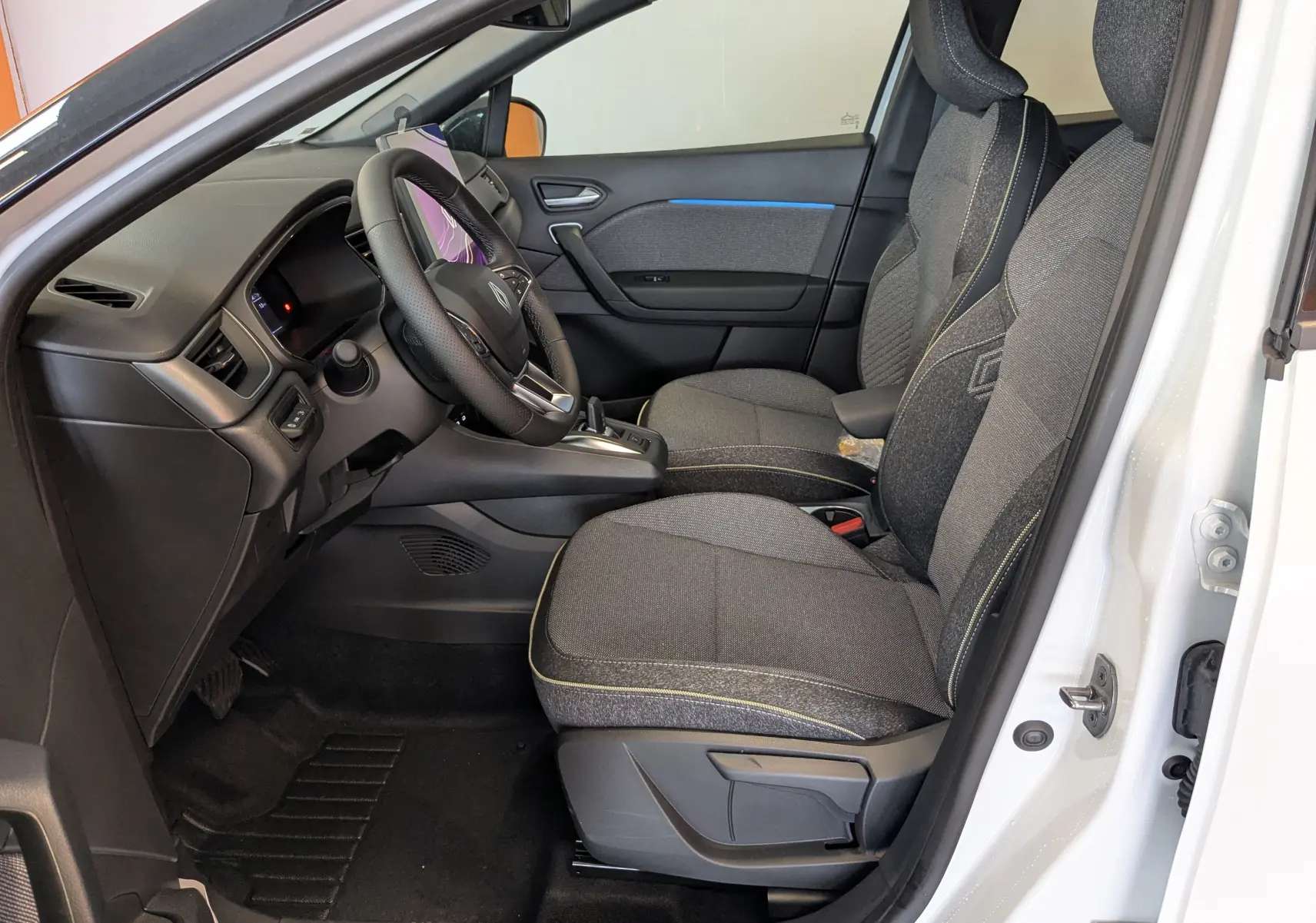 Intérieur côté conducteur du Renault Captur blanc nacré 2025, sièges tissu gris avec surpiqûres jaunes et tableau de bord moderne.