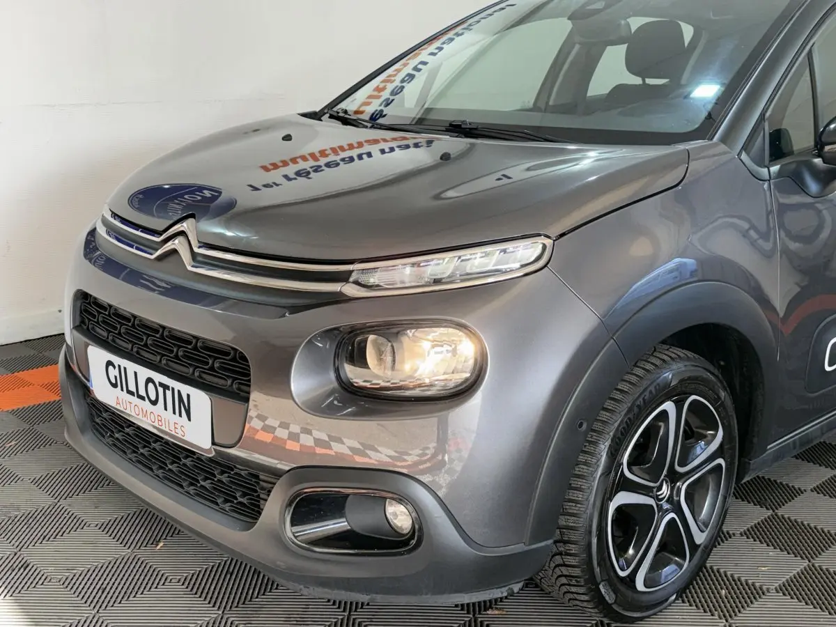 Vue 3/4 avant droit d'une Citroën C3 gris clair avec phares allumés et jantes alliage distinctives.