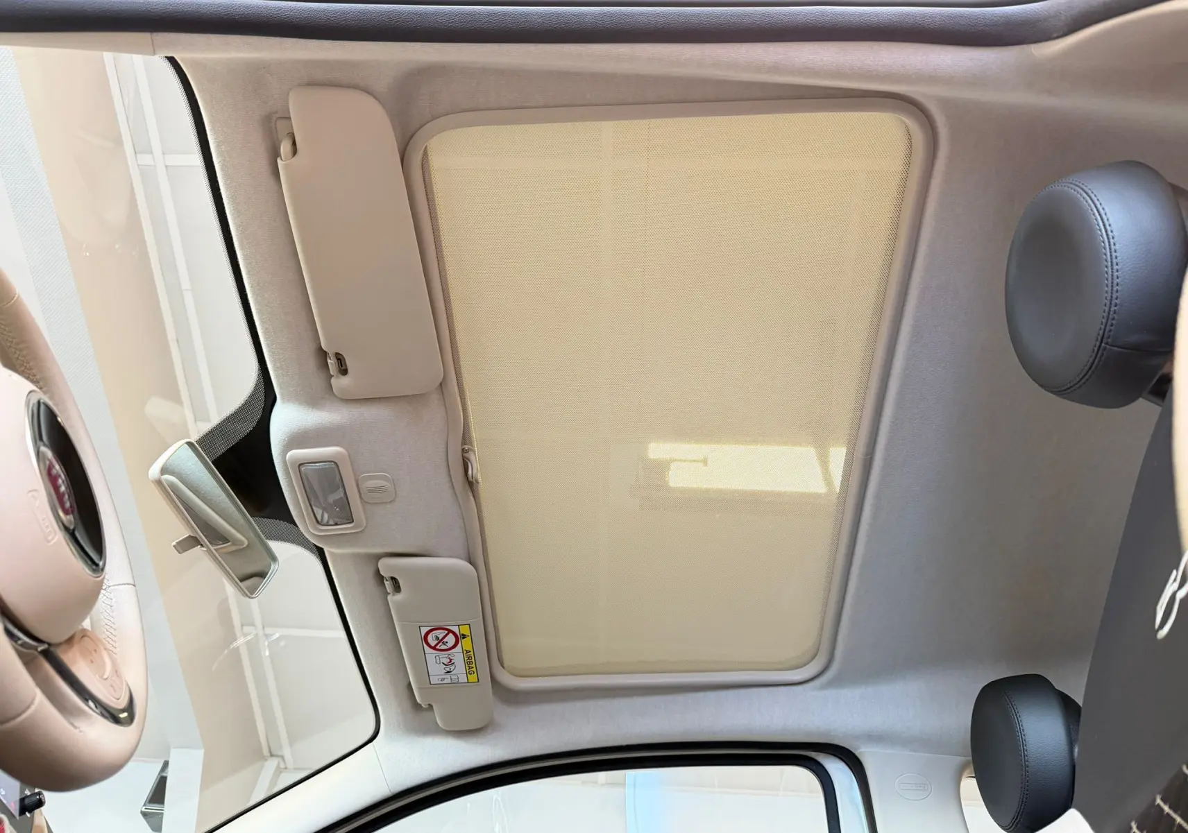 Vue intérieure du toit ouvrant avec pare-soleil beige de la Fiat 500 Dolcevita blanche, montrant le volant et appuie-tête noirs.