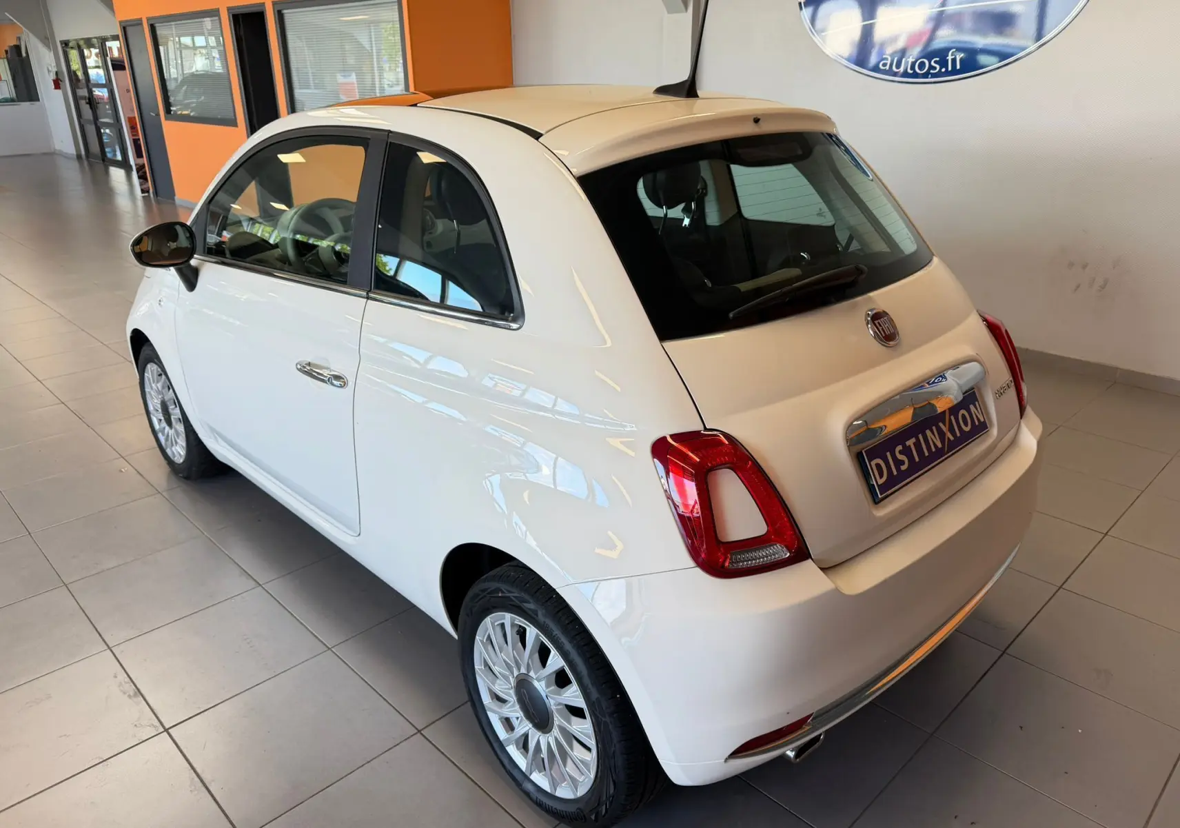 Vue 3/4 arrière droite d'une Fiat 500 blanche Bossa Nova dans un showroom avec jantes multibranches et logo distinctif.