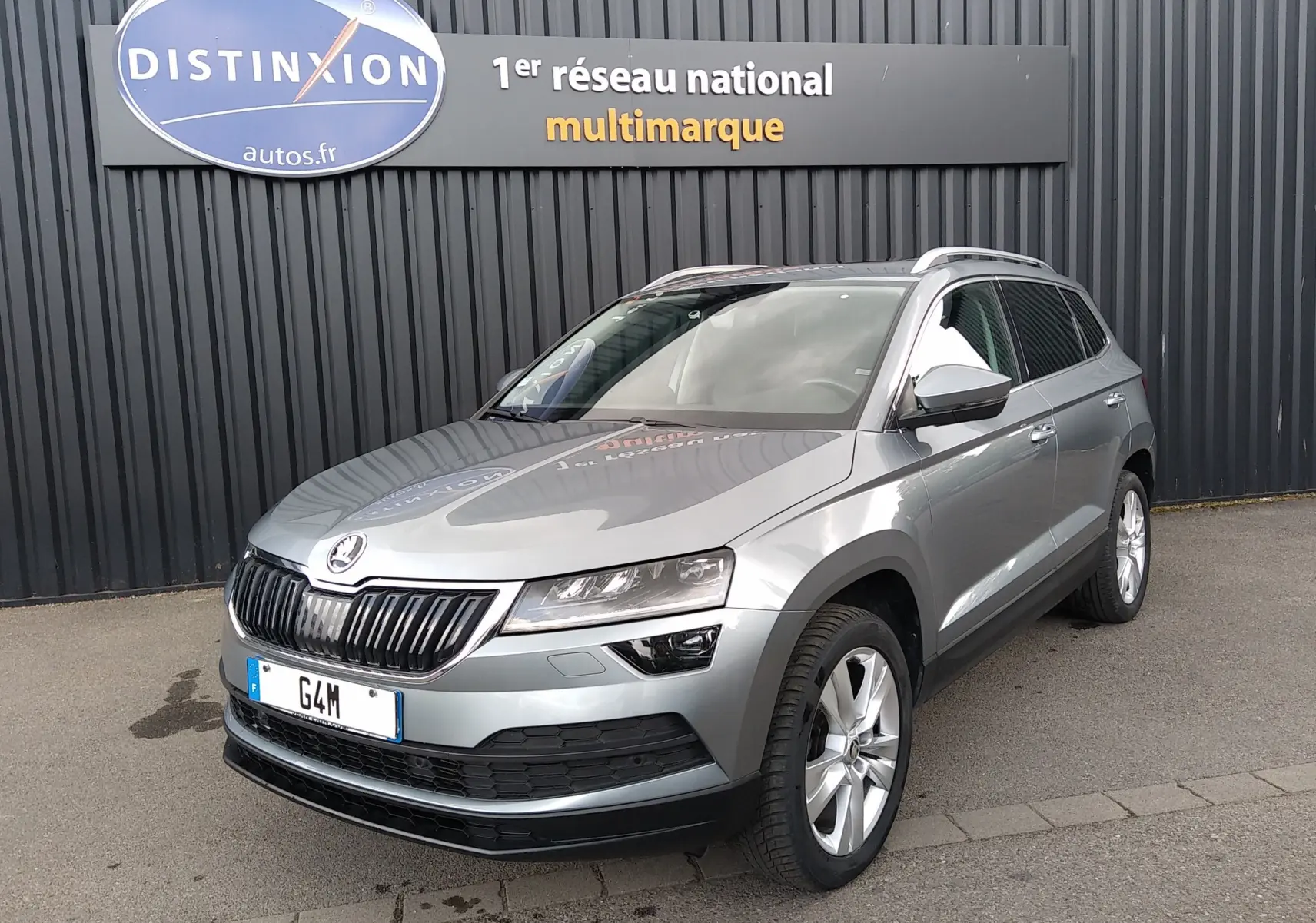 SUV Skoda Karoq gris en 3/4 avant droit, avec calandre noire et jantes alliage, stationné devant un mur noir.