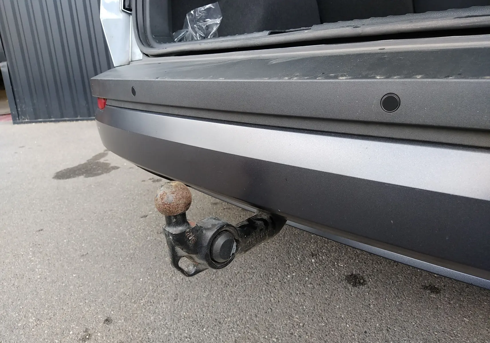 Vue rapprochée de l'attelage arrière rouillé d'un Skoda Karoq gris, coffre ouvert sur un sol goudronné.