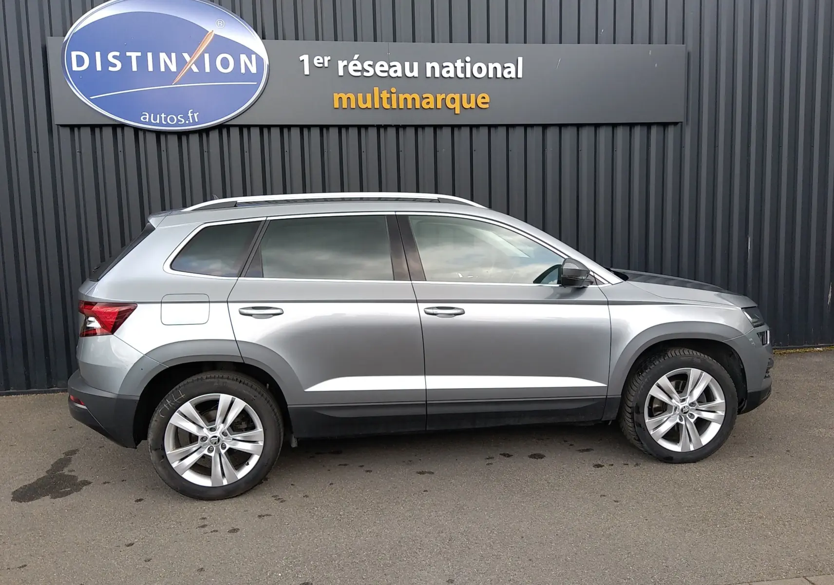 Vue de profil côté gauche d'un Skoda Karoq gris 2022 garé devant un mur noir avec un panneau Distinxion.