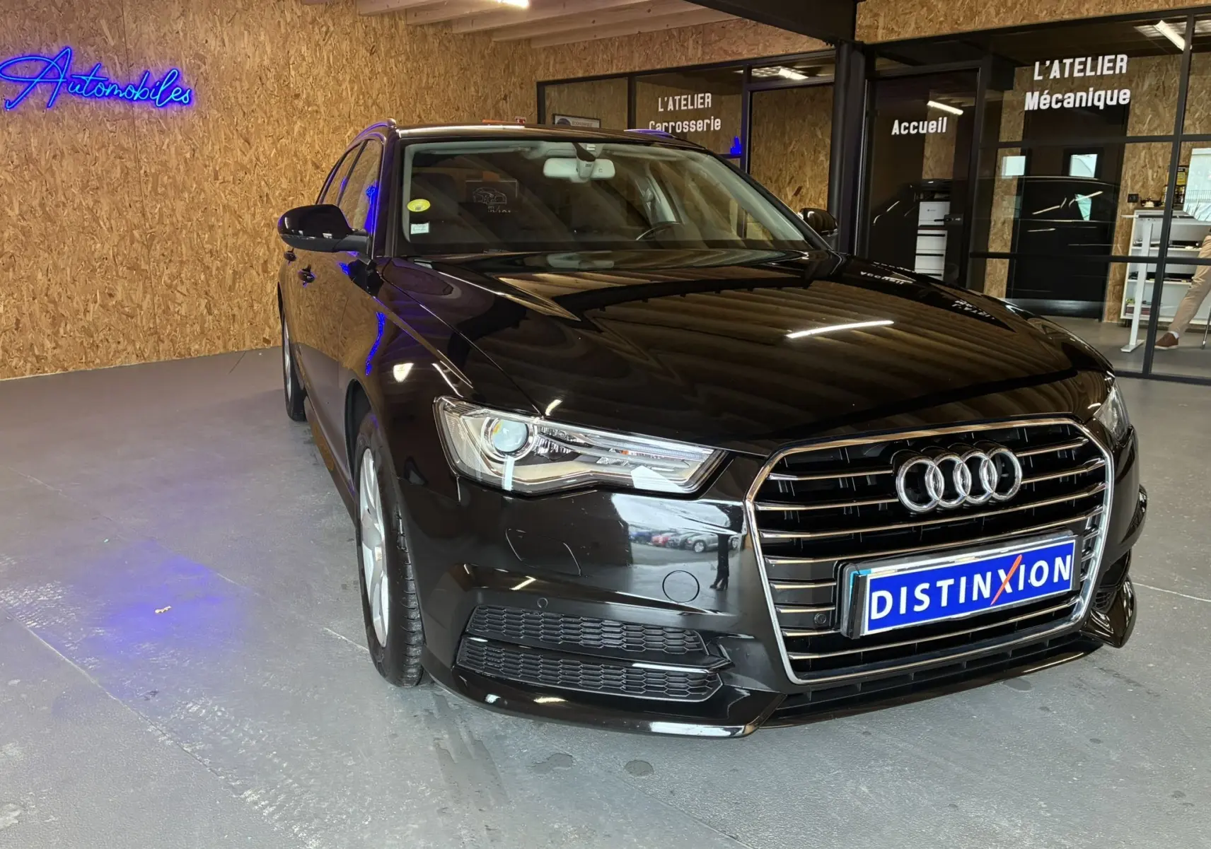 Audi A6 Avant noire vue de 3/4 avant droit dans un garage avec enseignes lumineuses en arrière-plan.