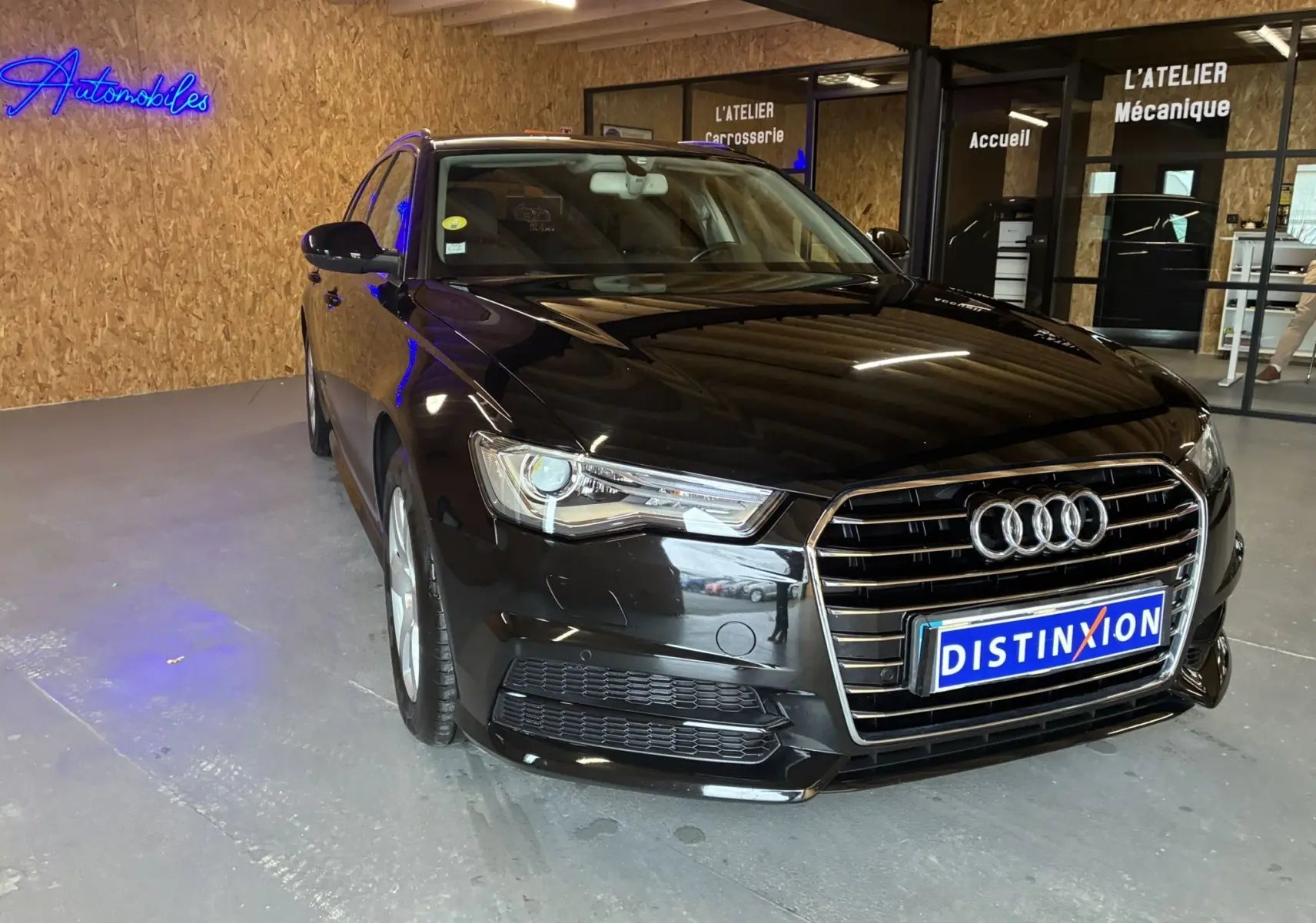 Audi A6 Avant noir vue 3/4 avant droit dans un garage avec enseignes lumineuses en arrière-plan.