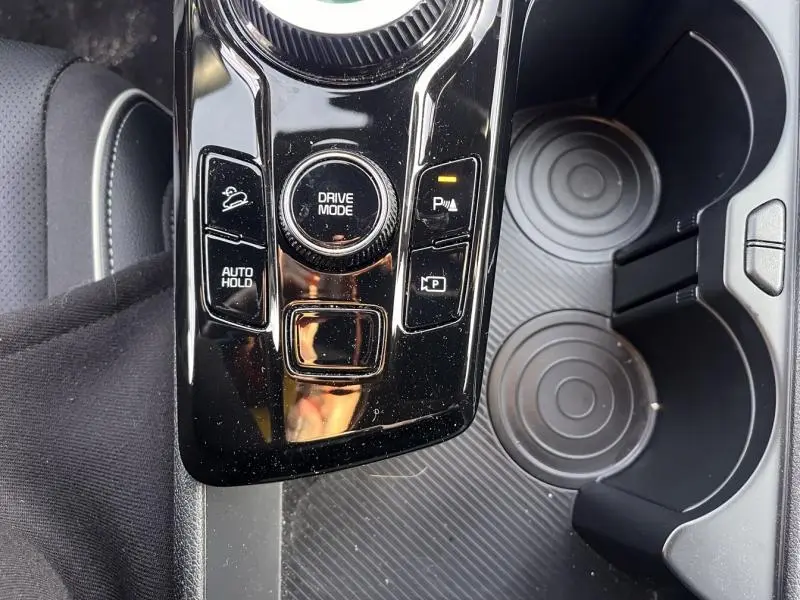 Vue rapprochée de la console centrale noire brillante du Kia Sportage blanc, avec commandes Drive Mode et porte-gobelets.