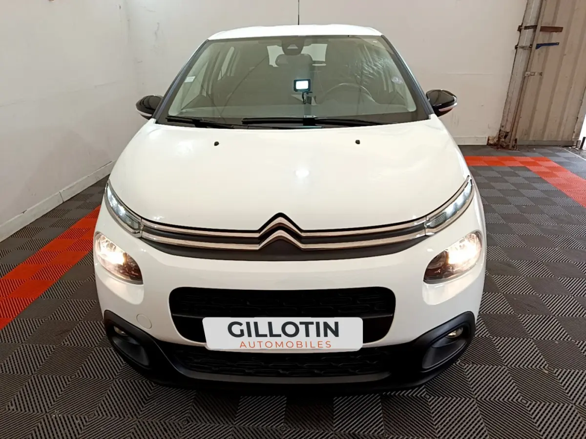 Citroën C3 blanche vue de face en intérieur, avec feux avant allumés et logo distinctif sur la calandre.