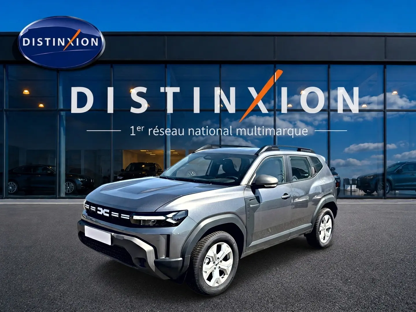 Dacia Duster gris schiste en 3/4 avant droit, avec barres de toit et jantes alu, devant une vitrine Distinxion.
