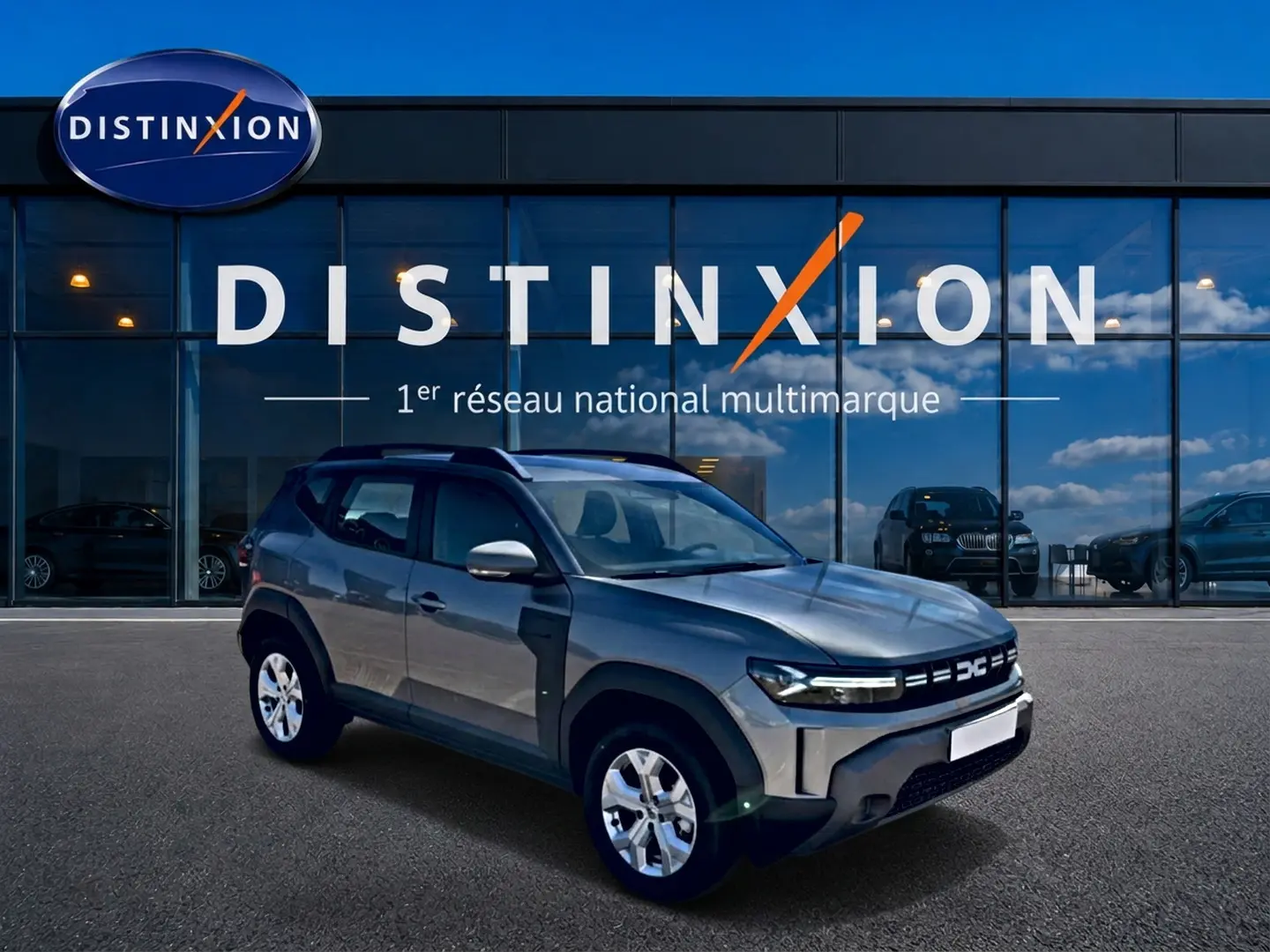 Dacia Duster gris schiste en 3/4 avant droit, avec jantes alu et barres de toit visibles devant vitrine Distinxion.