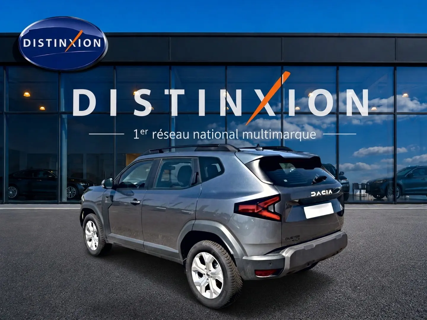 Vue 3/4 arrière droite du Dacia Duster gris schiste 2026 avec barres de toit et jantes alu devant un showroom Distinxion.