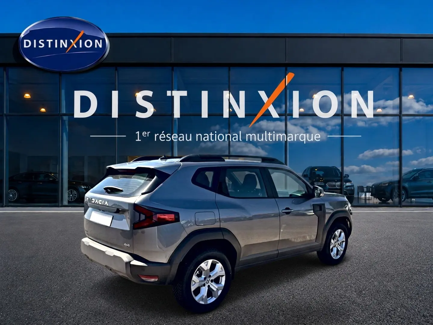 Vue 3/4 arrière droite d'un Dacia Duster gris schiste 2026 avec barres de toit, garé devant une vitrine Distinxion.