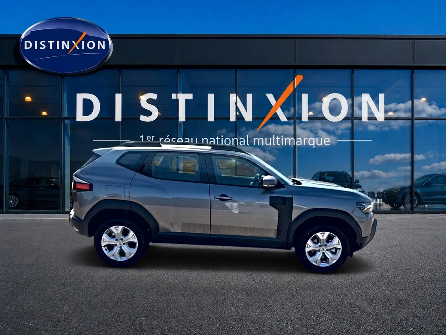 Vue latérale droite du Dacia Duster gris schiste 2026 devant une vitrine Distinxion, soulignant ses jantes alu et barres de toit.