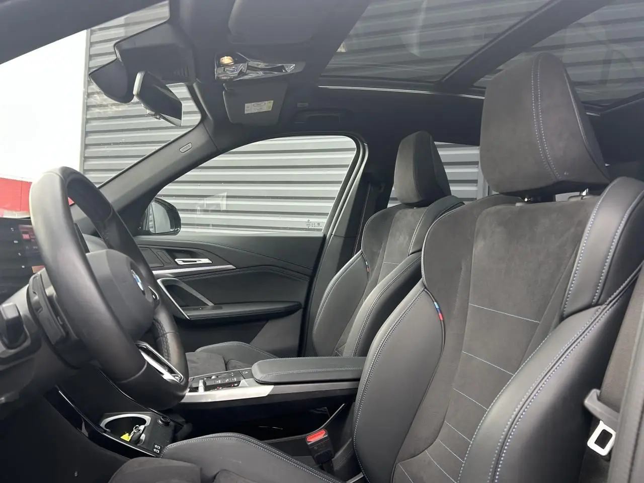 Intérieur BMW X1 2025 en vue côté gauche, sièges sport en alcantara noir avec surpiqûres bleues et toit panoramique.