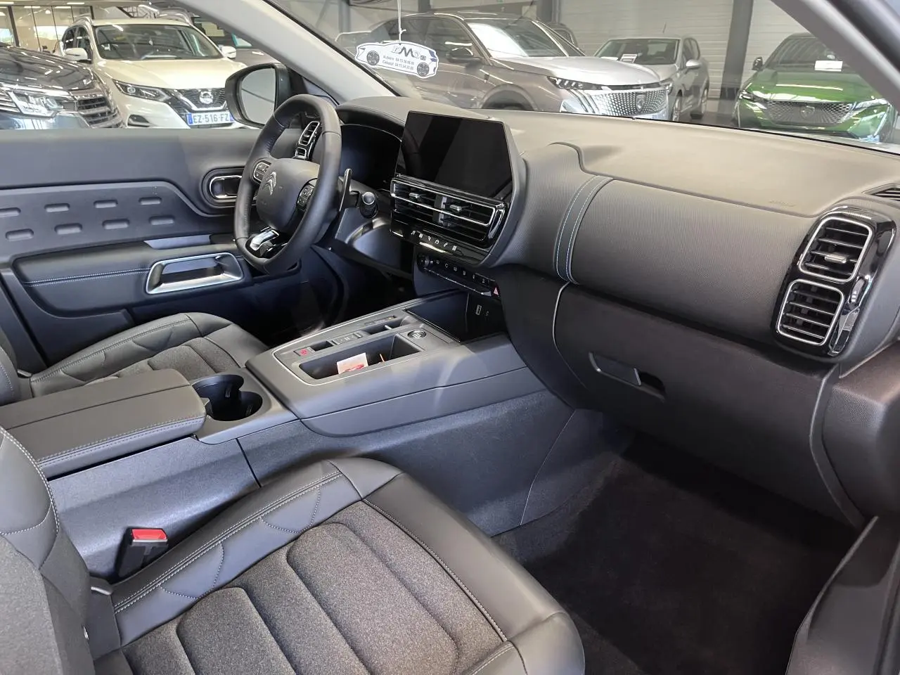 Intérieur avant du Citroën C5 Aircross 2025, vue côté conducteur, tableau de bord noir avec surpiqûres bleues et écran tactile central.