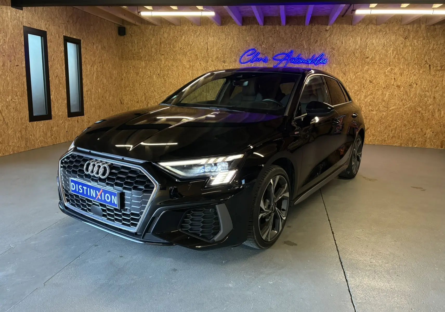 Audi A3 Sportback noir vue 3/4 avant droit en intérieur, avec calandre hexagonale et phares LED allumés.