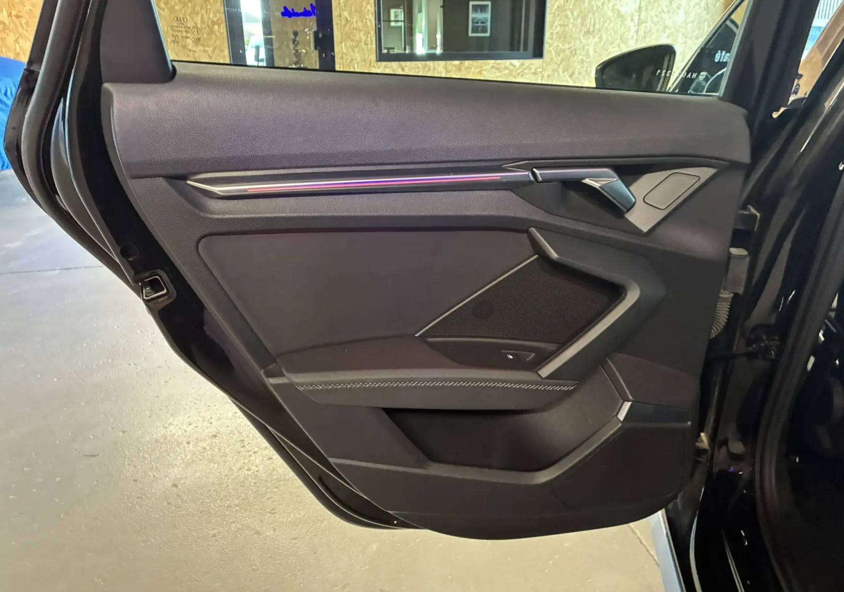 Porte arrière côté gauche ouverte d'une Audi A3 Sportback noire, mettant en valeur la garniture intérieure noire avec éclairage d'ambiance.