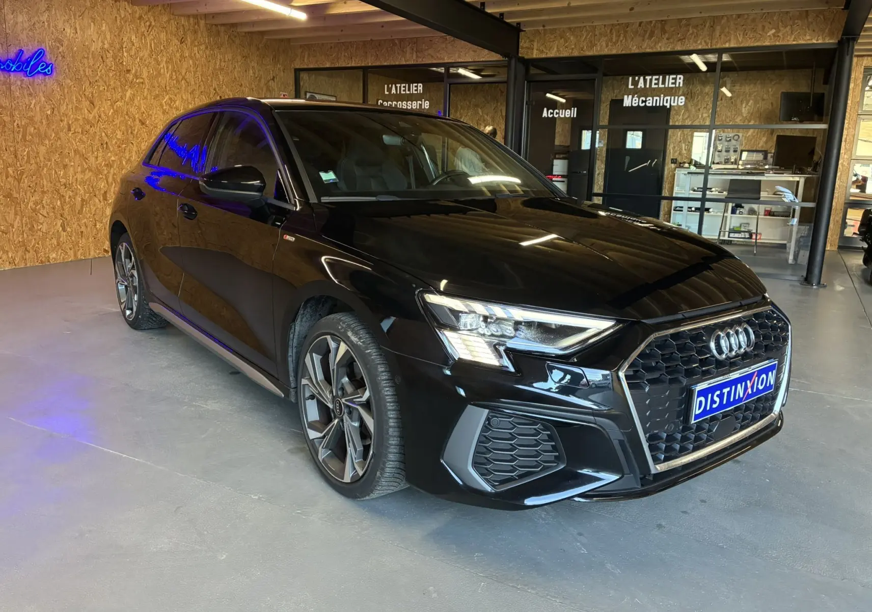 Audi A3 Sportback noir en 3/4 avant droit, avec calandre hexagonale et jantes alliage distinctives, stationnée en intérieur showroom.