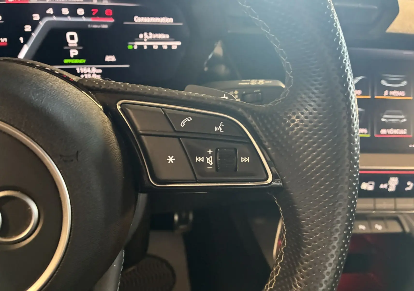 Gros plan sur les commandes du volant en cuir perforé noir d'une Audi A3 Sportback 2022 avec tableau de bord numérique en arrière-plan.