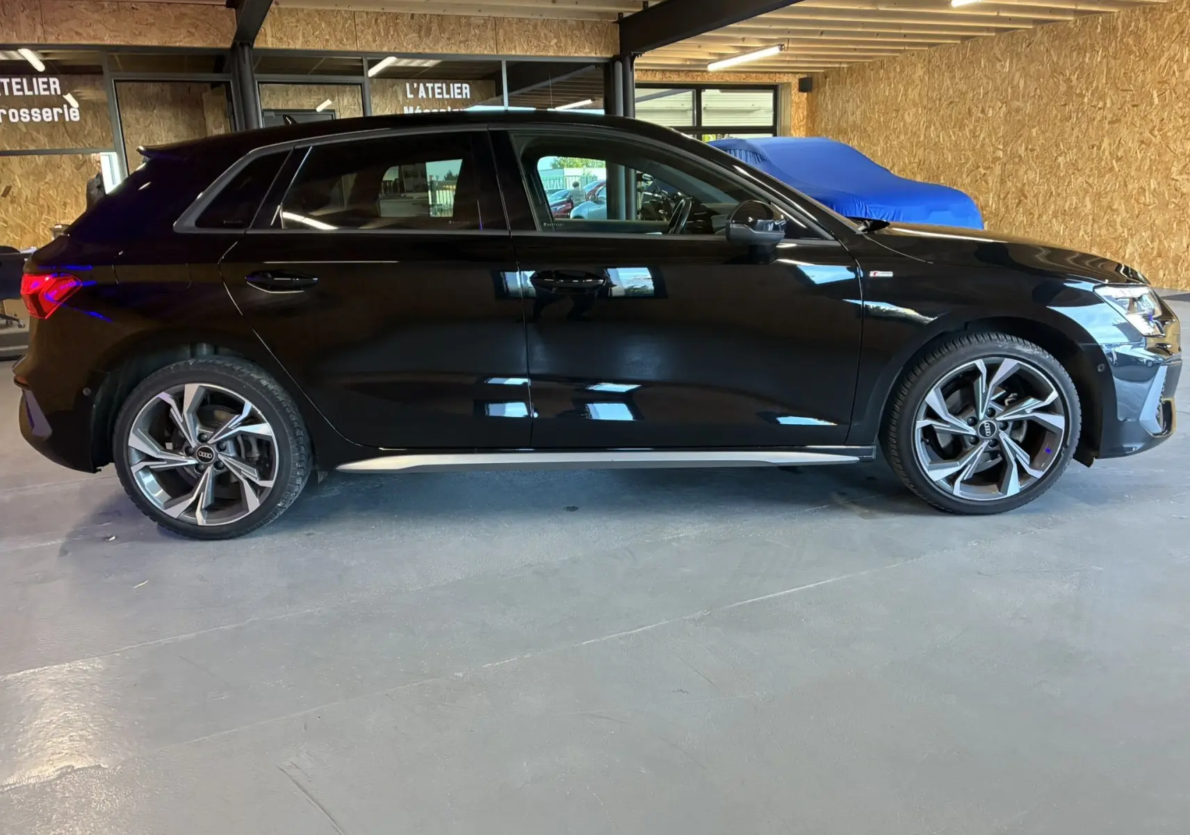 Profil côté gauche d'une Audi A3 Sportback noire 2022 S Line avec jantes alliage et intérieur cuir noir visible en atelier.