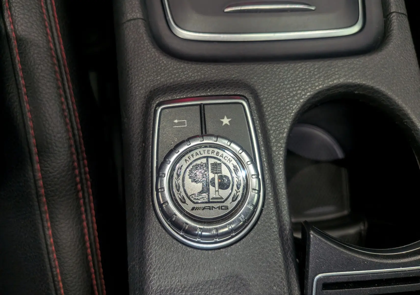 Gros plan sur la molette AMG en aluminium brossé avec logo Affalterbach, entourée de cuir noir à surpiqûres rouges.