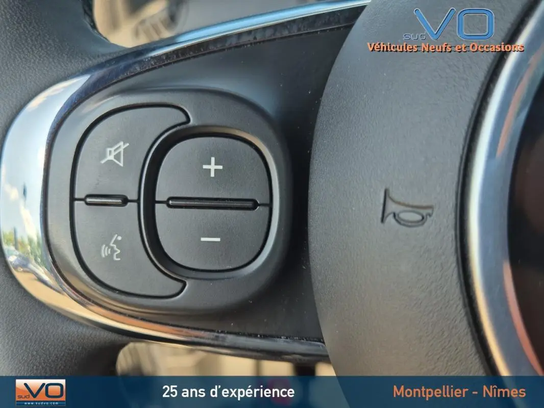 Gros plan sur les commandes de volume au volant d'une FIAT 500C noire 2024 avec roue de secours visible.