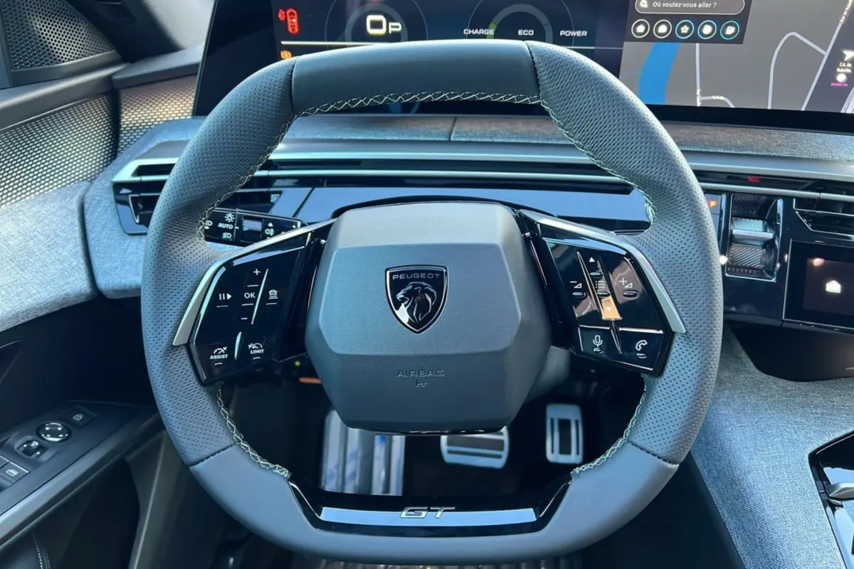 Vue rapprochée du volant cuir perforé gris du Peugeot 3008 Hybrid GT avec logo lion et inscription GT, tableau de bord digital en arrière-plan.
