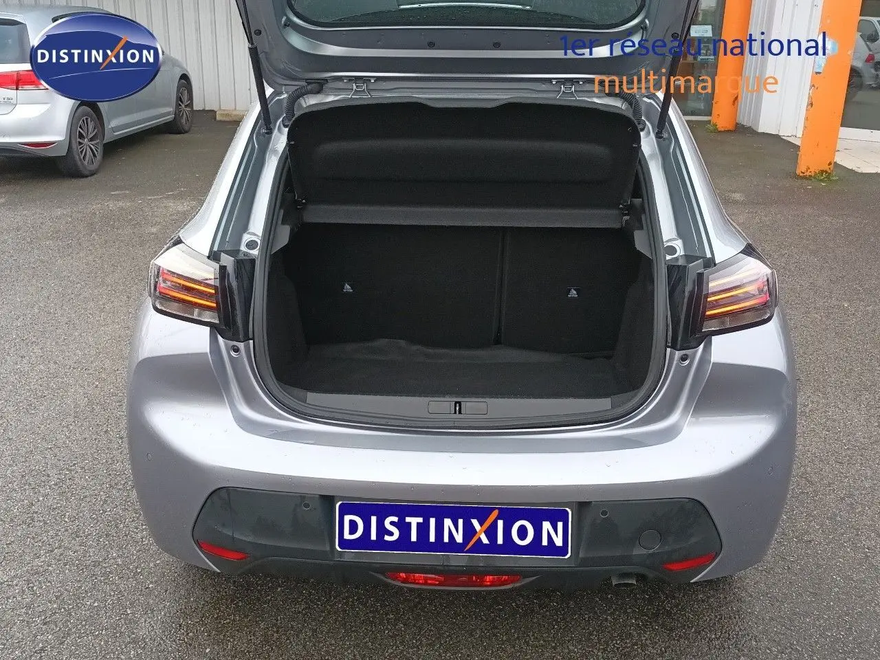 Vue arrière ouverte sur le coffre d'une Peugeot 208 gris Artense métal, avec feux arrière allumés et plaque Distinxion.