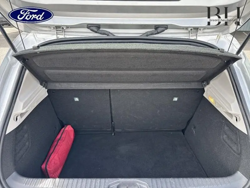 Coffre ouvert vu de l'arrière d'une Renault Clio gris platine avec un sac de secours rouge posé à gauche.