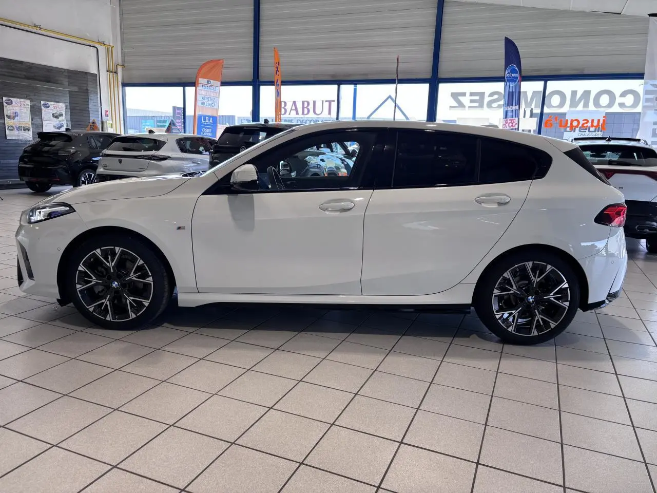 BMW Série 1 120d M Sport blanc vue de profil côté gauche en intérieur, avec jantes alliage noires et vitres teintées.