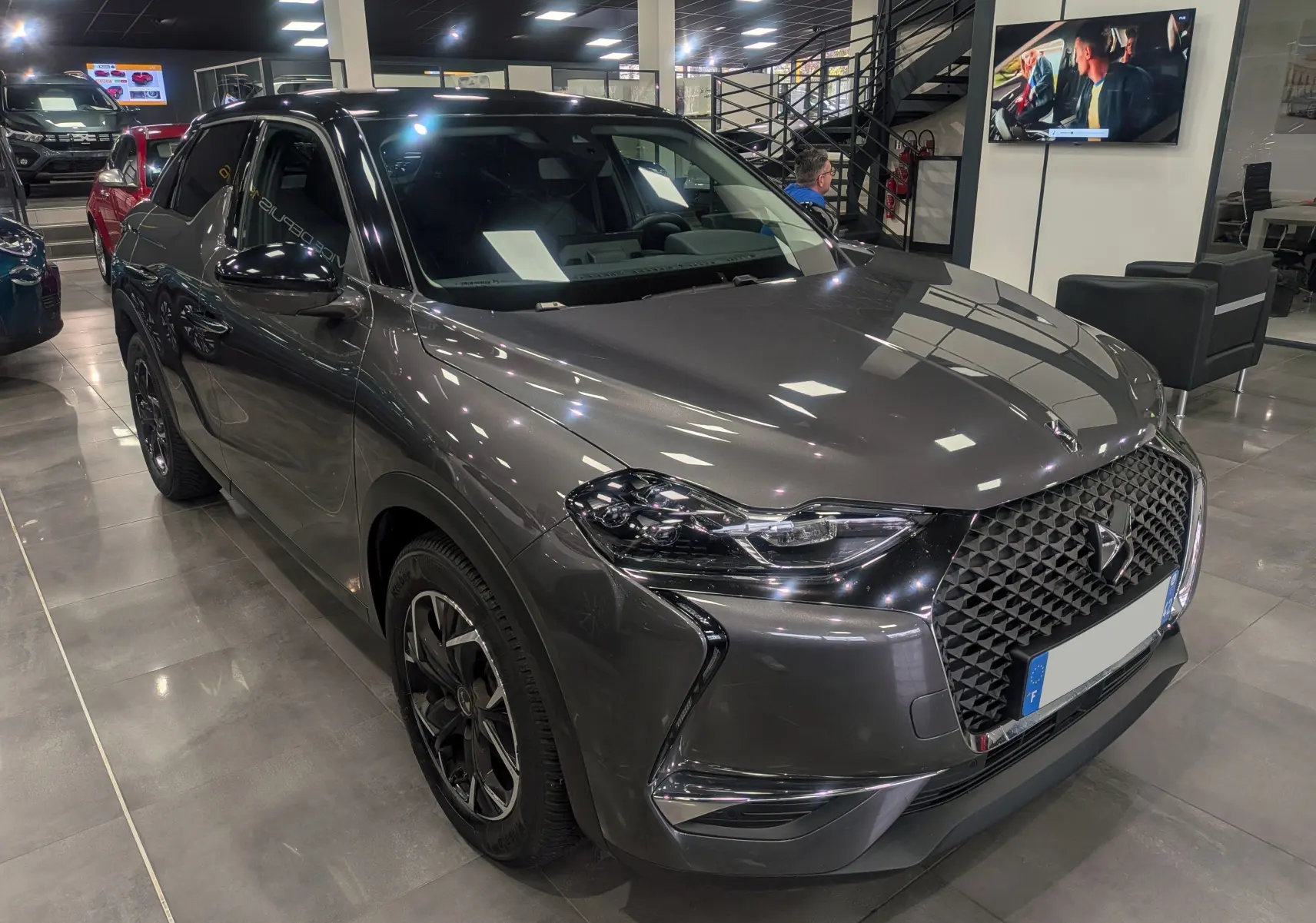 Vue 3/4 avant droit d'un DS3 Crossback gris Platinium avec toit noir et calandre distinctive en losange dans un showroom.