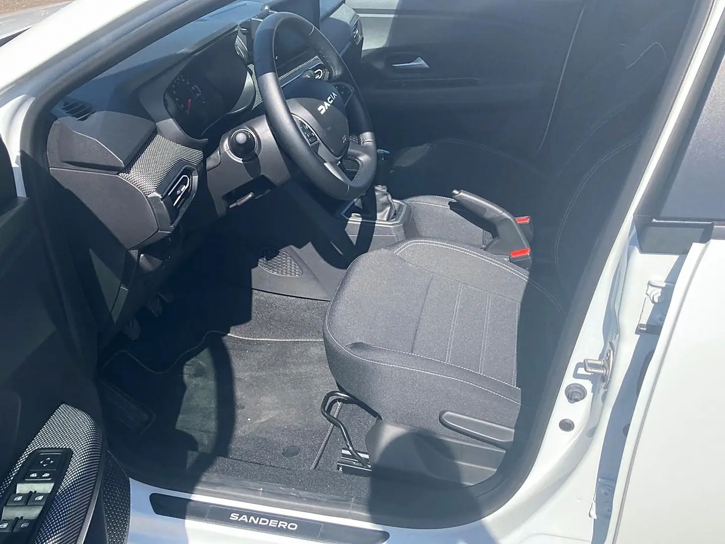 Vue intérieure côté conducteur d'une Dacia Sandero blanche 2025, volant multifonctions et sièges tissu gris.