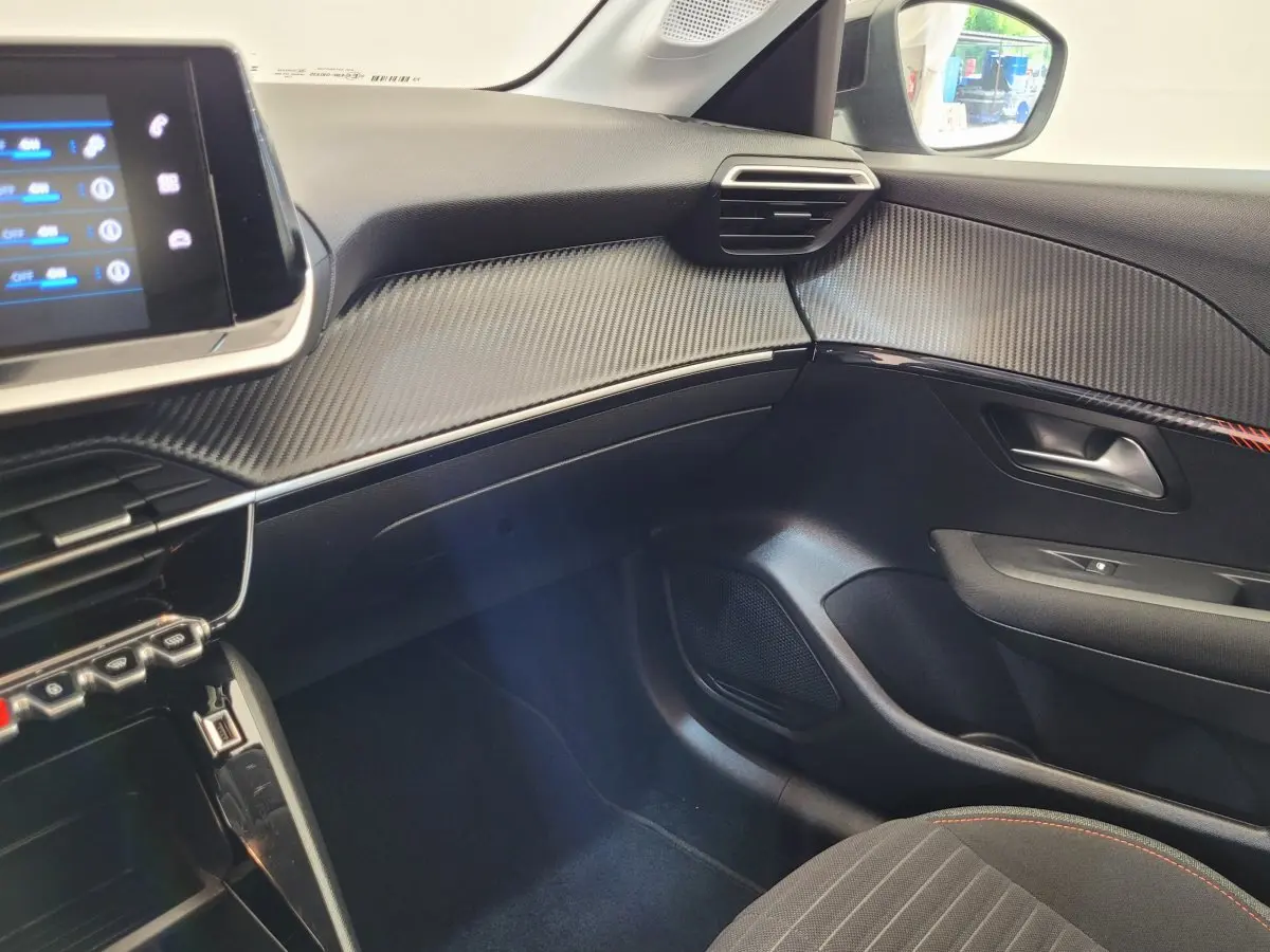 Vue intérieure côté passager du tableau de bord et porte avec finition noire et détails en fibre de carbone du Peugeot 208 2023.