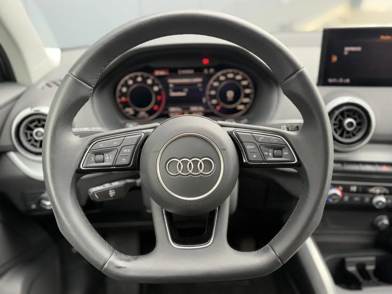 Vue rapprochée du volant cuir multifonction de l'Audi Q2 30 TFSI S-Line 2024 avec tableau de bord numérique en arrière-plan.