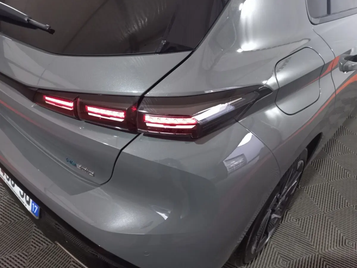 Vue 3/4 arrière droite d'une Peugeot 308 PHEV 180 gris foncé avec feux arrière LED allumés et prise de recharge visible.