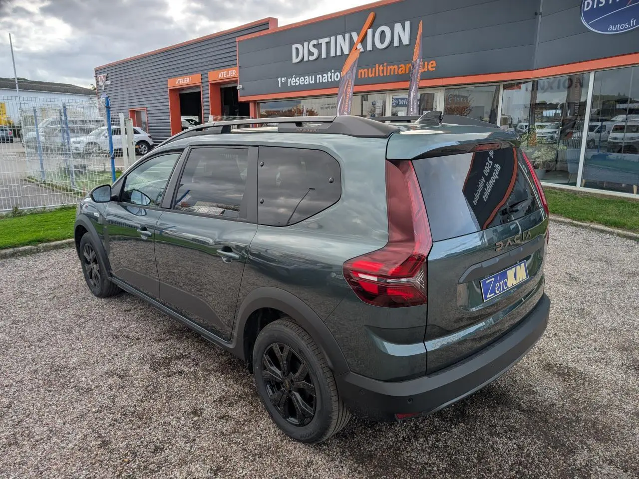 Vue 3/4 arrière droite d'un Dacia Jogger vert métallisé avec jantes noires et barres de toit sur un parking.