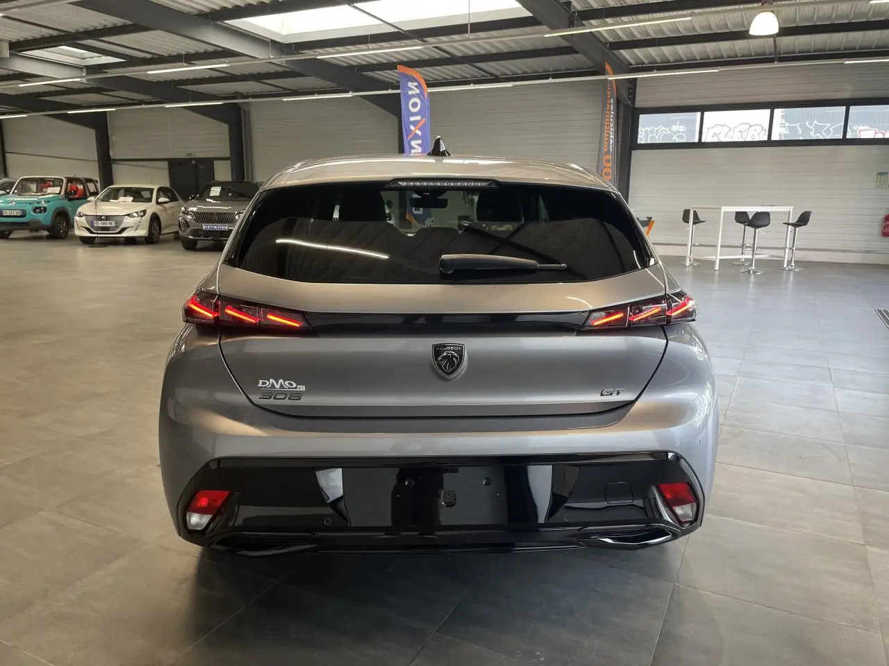 Vue arrière d'une Peugeot 308 GT gris Artense avec feux arrière LED 3 griffes et bandeau noir brillant en intérieur showroom.