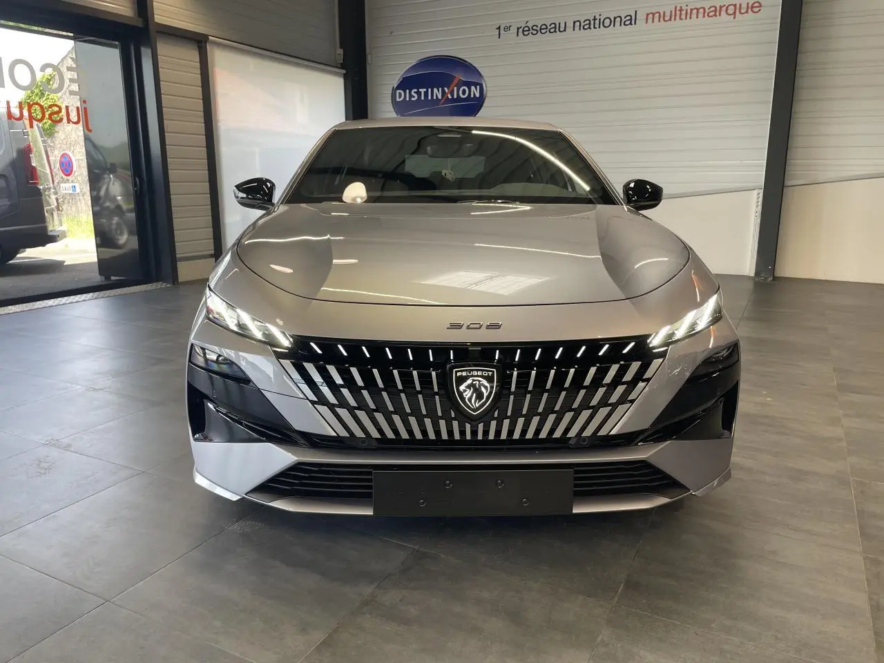 Vue frontale d'une Peugeot 308 gris Artense avec calandre noire brillante et logo lion lumineux en showroom.