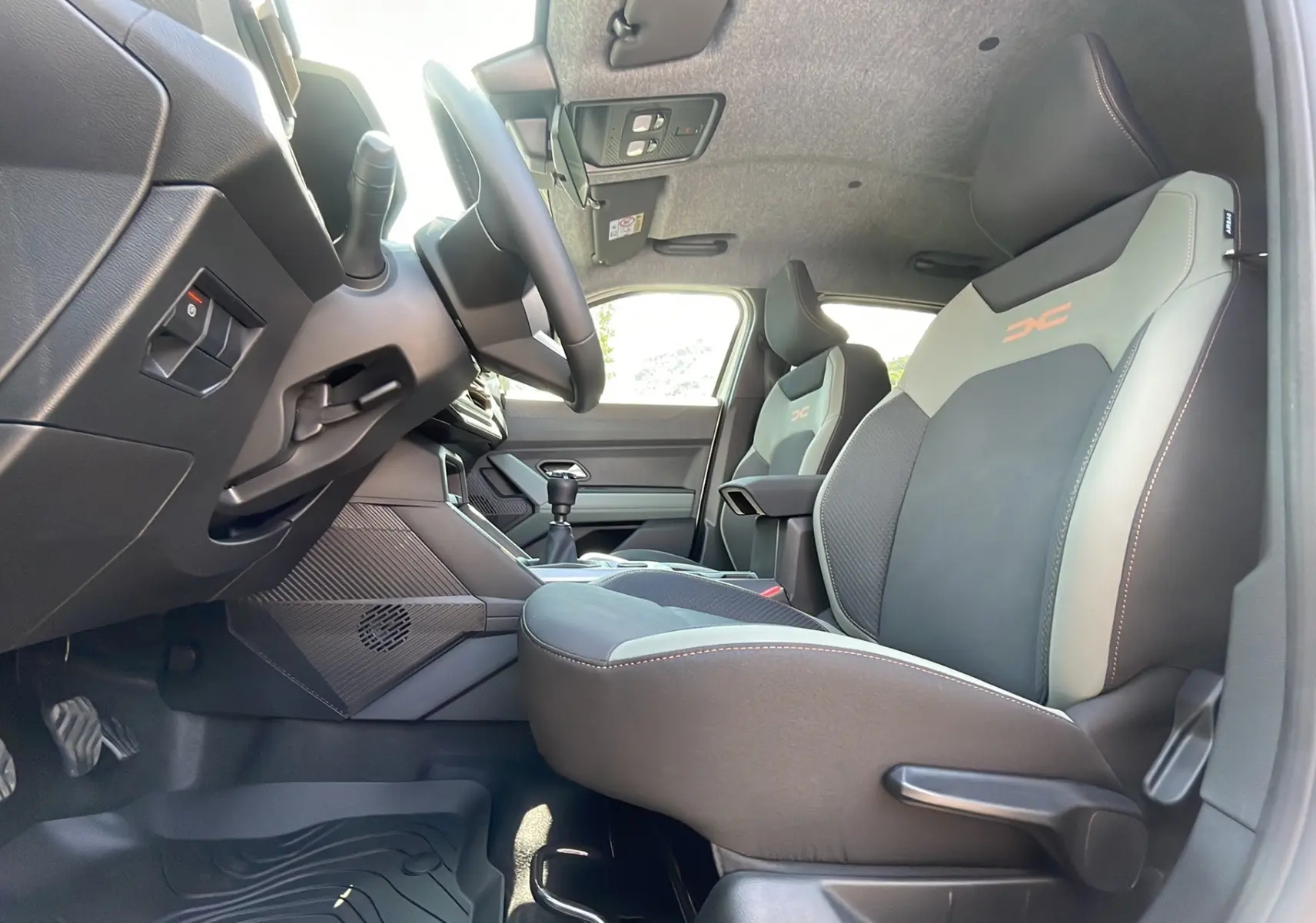 Vue intérieure côté conducteur du Dacia Duster 2025 avec sièges sport gris et logo orange, volant et levier de vitesse manuelle visibles.
