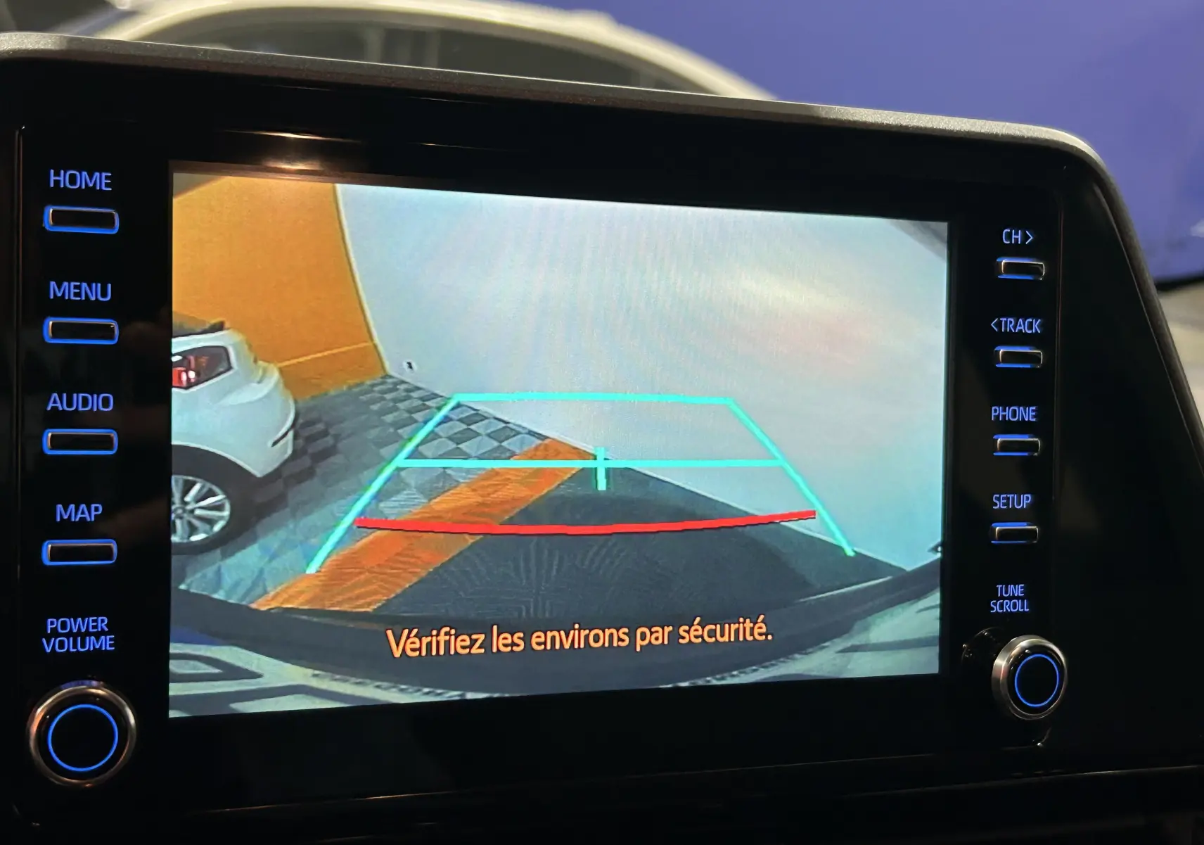 Écran de caméra de recul montrant le sol et un mur orange, avec une voiture blanche garée à gauche.