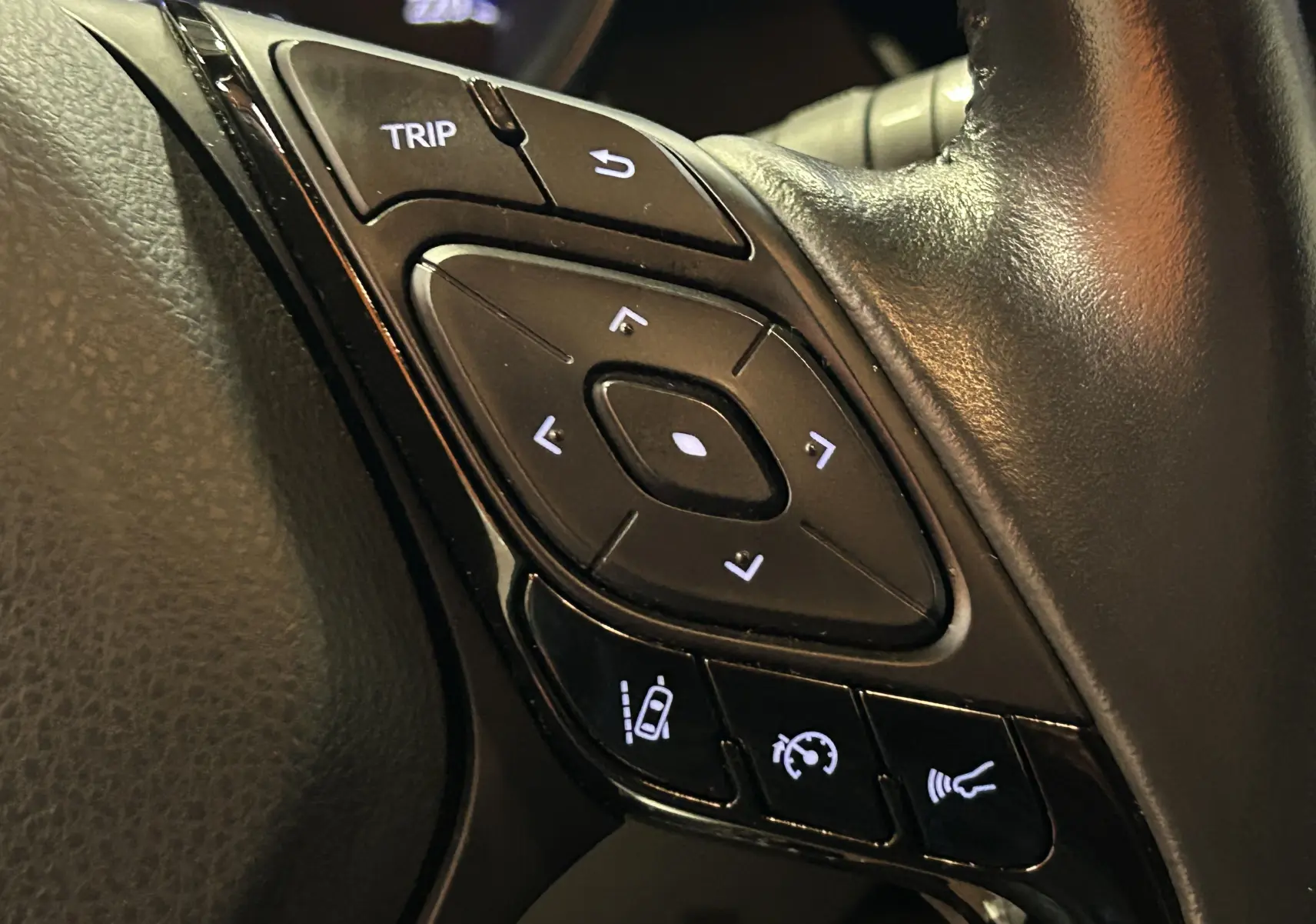 Gros plan sur les commandes du volant en cuir noir du Toyota C-HR Hybride 122h Edition 2WD, boutons rétroéclairés blancs.
